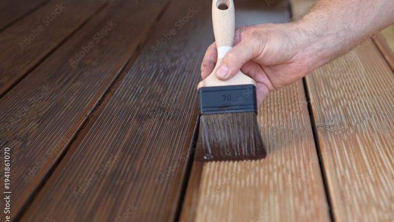 Eine Hand hält einen Pinsel und streicht braune Farbe auf Holz.